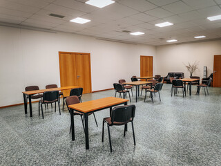 A spacious conference room with wooden tables and black chairs arranged for meetings or events. The minimalist interior features plain walls, wooden doors, and overhead lighting.