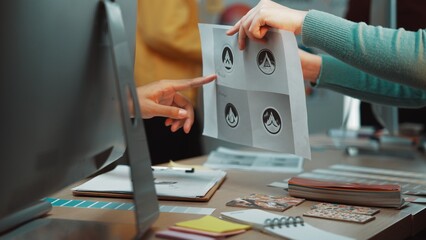 Two creative professionals engage in a collaborative discussion about logo designs. They review printed graphics and share ideas in a bright and modern office environment. SACTR