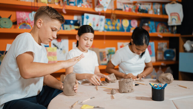 Caucasian smart teenager modeling clay while craving glass of clay at art lesson. Group of multicultural happy student working at dough in pottery workshop. Creative activity concept. Edification.