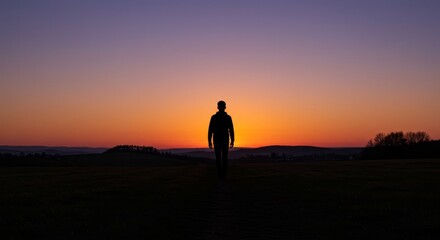 Fototapeta premium Silhouette of Person at Sunset - Solitary figure walking towards a vibrant sunset over a tranquil landscape. Peaceful and serene scene