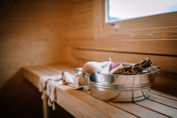 A cozy sauna interior with a wooden bench, a metal bucket, a wooden ladle, a sauna whisk, and a linen towel, creating a relaxing ambiance.