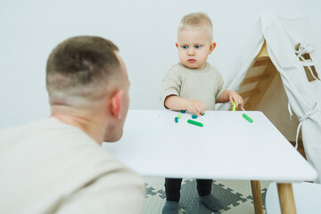 A little one-year-old boy is sculpting plasticine while standing at the table. The child is playing...