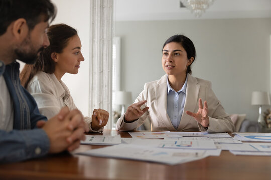 Indian financial broker woman and Caucasian couple of customers discussing investment strategy, planning savings, convenient taxes payment, talking at table with paper analytic diagrams at home