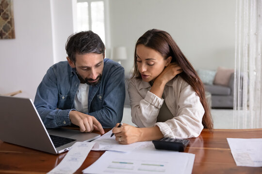 Serious young middle aged couple analyzing personal domestic budget at home, checking paper bills, receipts for payment, discussing expenses at table with laptop and calculator