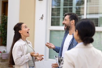 Fototapeta premium Cheerful excited couple of new homeowners celebrating buying home, throwing and catching keys from new accommodations, dwelling, standing with realtor outside of house, having fun