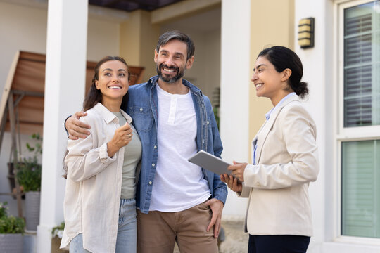 Positive couple of new homeowners and young Indian realtor woman standing outside of house, looking away, discussing apartment buying deal. Looking away, viewing backyard, neighborhood - Powered by Adobe