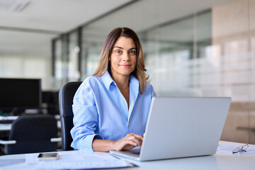 Happy smiling mature professional business woman of middle age, bank worker, hr manager, corporate executive or company worker looking at camera working on laptop computer in office, portrait.
