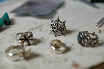 Close-up of various silver jewelry pieces, including a spider web design, arranged on a workshop table under soft lighting. Detailed shots showcase craftsmanship