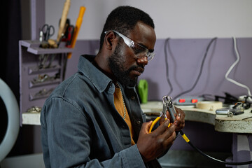 Portrait of African American man in workshop environment wearing safety glasses and using pliers...