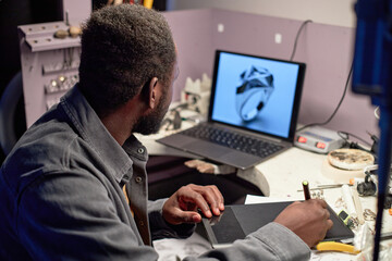 African American man working on jewelry design using computer displaying intricate ring design. Engaging in creative process with various tools and equipment in organized workspace