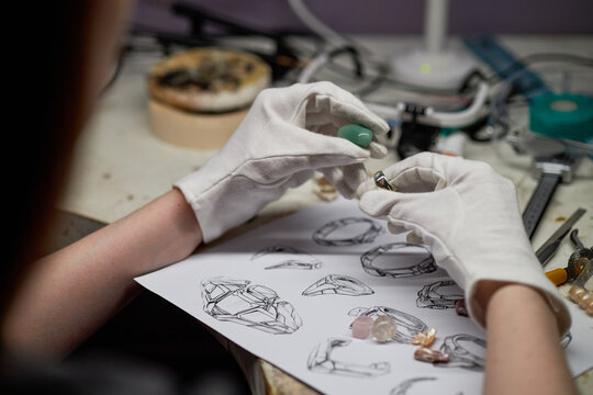 Carefully handling gemstone with gloved hands, focusing on jewelry design details including various ring sketches on paper, workbench filled with tools
