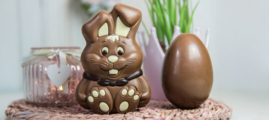 A chocolate bunny and a chocolate egg stand on a table decorated with hyacinths and candles. Blurred pink decorative elements.