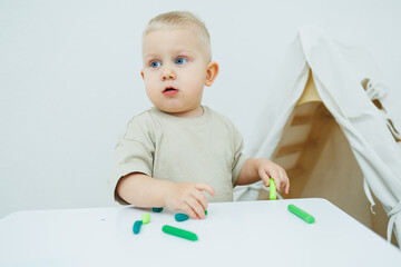 A little one-year-old boy is sculpting plasticine while standing at the table. The child is playing...