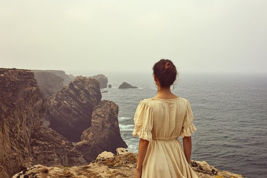Woman in a beige dress gazes at a misty ocean and dramatic cliffs.