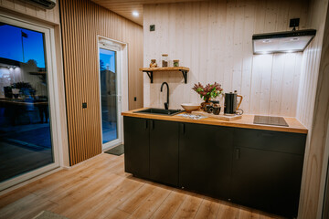 A modern kitchen with black cabinets, wooden countertops, and warm lighting, featuring a sink, stove, open shelving, and cozy decor.