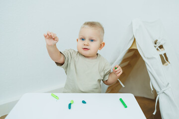 A little one-year-old boy is sculpting plasticine while standing at the table. The child is playing...