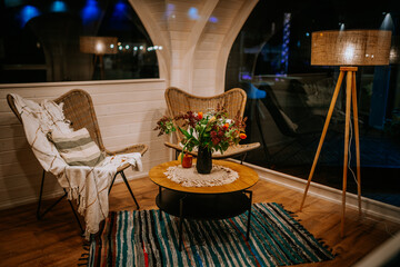 A cozy indoor lounge with wicker chairs, a wooden table, a floral centerpiece, a floor lamp, and warm lighting, creating a relaxing atmosphere.