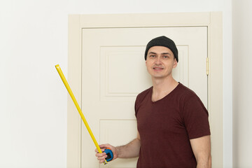 A man stands beside a closed door, holding a yellow measuring tool, dressed in a casual outfit. He...
