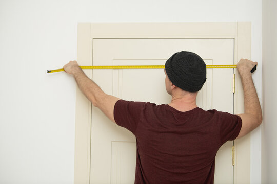 A man in a black beanie takes precise measurements of a door frame using a measuring tape. The task takes place indoors during bright daylight