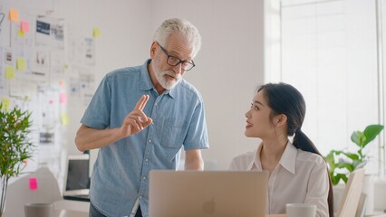 Fototapeta premium Mentoring in modern office: Senior man explains project to Asian woman at computer.