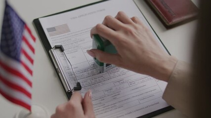 Hands of female consular officer examining and approving visa application form with official stamp at table with American flag and passport on it. Close-up view