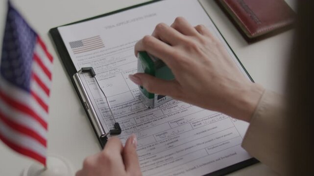 Hands of female consular officer examining and approving visa application form with official stamp at table with American flag and passport on it. Close-up view