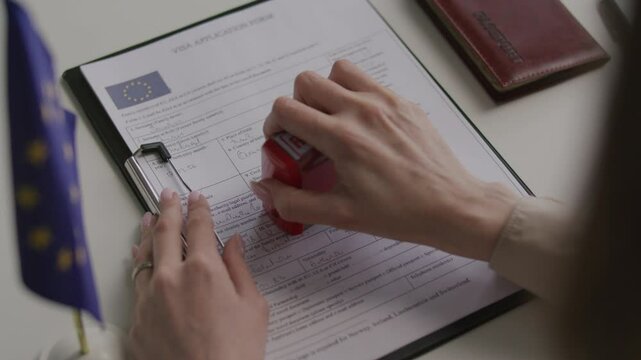 Hands of female consular officer pressing red rejected stamp on Schengen visa application form with EU flag and passport beside it on office desk. Close-up shot