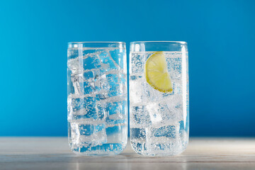 Refreshing soda water with ice cubes and slice of lemon in glasses on wooden table against light blue background, closeup