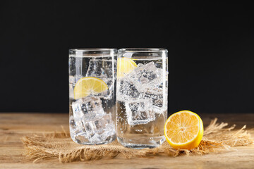 Refreshing soda water with ice cubes in glasses and cut lemon on wooden table against black background, closeup