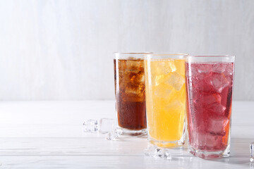 Soda water of different flavors in glasses with ice cubes on white wooden table, space for text