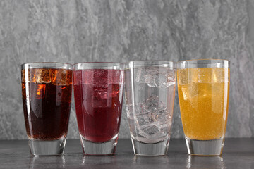 Sweet soda water of different flavors with ice cubes in glasses on grey table
