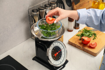 Woman adding tomato into bowl on mechanical kitchen scale at white marble countertop indoors,...