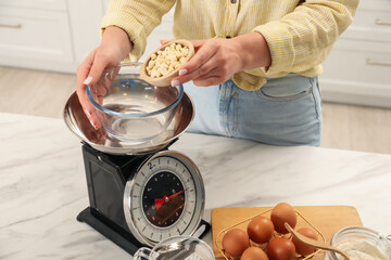 Woman adding peanuts into bowl on mechanical kitchen scale at white marble table indoors, closeup