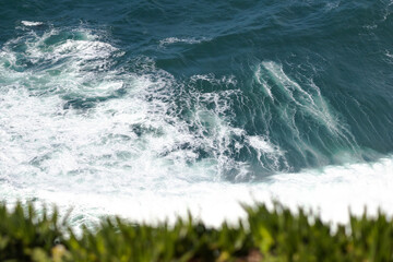 Fototapeta premium Rocky Cape Roca in Sintra cliffs rise steeply from the Atlantic Ocean, covered with patches of greenery. Waves crash against the jagged shoreline, creating white foam contrasting.
