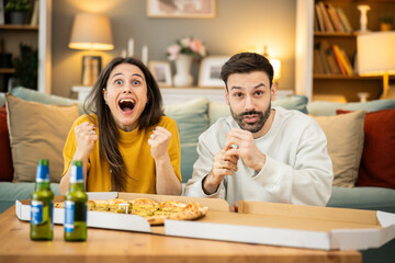 A joyful couple celebrating with pizza and drinks, showing excitement and happiness in their cozy living room setting.