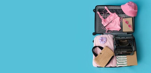 Open suitcase with vintage typewriter and travel accessories on light blue background with space...