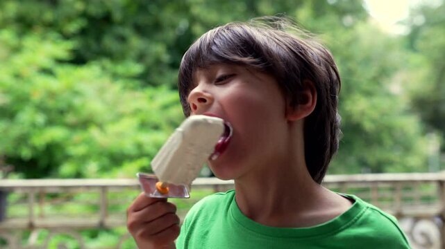 Young boy enjoying an ice cream outdoors, close-up of his face as he savors a cold treat on a warm day in a natural, green setting. Joyful moment of a child indulging in a summer snack