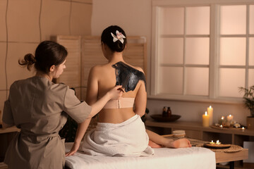 Naklejka premium Young woman undergoing mud treatment in dark spa salon, back view