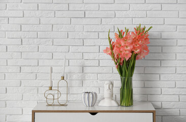 Vase with gladiolus flowers, Moai statue and candles on commode near white brick wall