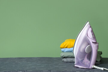 Board with electric iron and stack of clothes near green wall in room