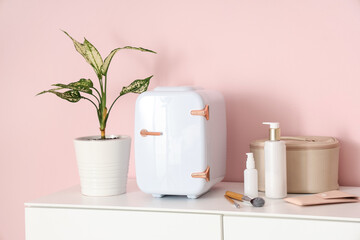 Small cosmetic refrigerator, houseplant and cosmetic bag on chest of drawers against pink background. Closeup