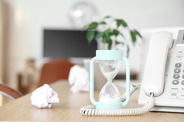 Hourglass with telephone and crumpled paper on desk in office, closeup