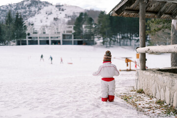 Small child stands near the gazebo on a snowy hill and looks at the skating rink with lugers. Back view