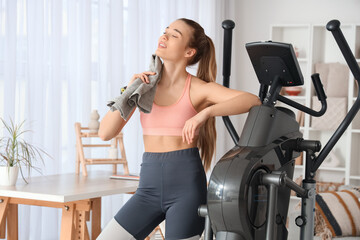 Fototapeta premium Sporty young woman with towel near modern exercising bike at home