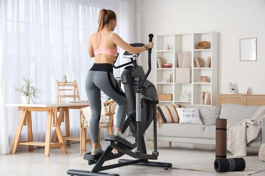 Sporty young woman training on modern exercising bike at home, back view