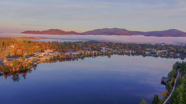 A stunning 4K drone shot of a minor lake in Lake Placid, Ithaca, NY, surrounded by autumn trees, houses, mountains, and clouds, taken during sunset.