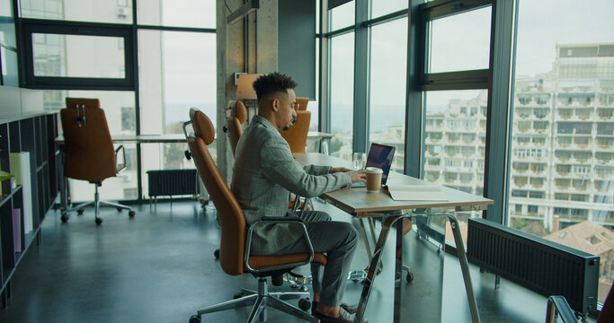 A businessman working on a laptop in a modern, open office with large windows and city views, showcasing a professional and productive workspace.