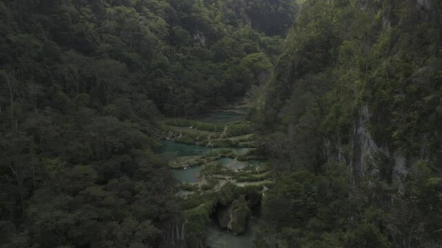 Drone lowers in front of waterfalls from pools over the Cahabon River, as a cloud passes overhead casting a shadow on the valley in Semuc Champey, Guatemala