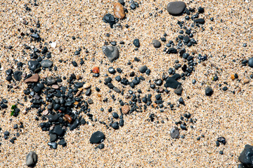 A close-up view of textured beach sand dotted with various dark pebbles and smooth stones, capturing the intriguing natural patterns and formations found by the shore.