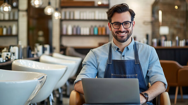 Male hairstylist wearing apron and glasses working on laptop in modern hair salon. Professional barber sitting in chair with smile. Business and entrepreneurship in beauty industry